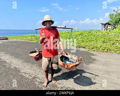 Kusamba, Indonésie. 16 février 2024. Le producteur de sel Nyoman Warta sur la plage de Kusamba. Il utilise des conteneurs simples pour transporter de l'eau de mer à terre tous les jours. Les touristes peuvent acheter le sel extrait naturellement et enrichi en minéraux directement sur place. Crédit : Carola Frentzen/dpa/Alamy Live News Banque D'Images