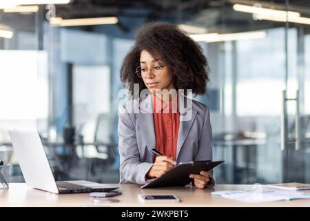 Femme focalisée dans des lunettes rondes regardant l'écran du pc tout en remplissant le document sur le tableau d'écriture dans l'intérieur du bureau. Entrepreneur confiant copiant des informations de courrier électronique à des papiers imprimés. Banque D'Images