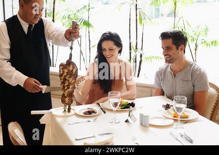 Couple heureux, date et serveur avec kebab de viande pour servir, dîner fin ou dîner romantique à table. Jeune homme et femme appréciant la brochette de coupe de chef Banque D'Images