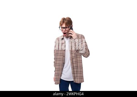 jeune homme caucasien avec les cheveux roux dans des lunettes et une chemise à carreaux parle soigneusement au téléphone et est surpris Banque D'Images