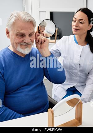 L'homme mature avec l'aide auditive ITE se regarde dans le miroir tenu par l'audiologiste souriant, et essaie l'appareil auditif. Appareil auditif élégant Banque D'Images