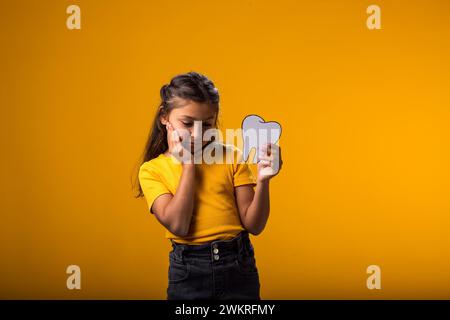 Enfant fille tenant une dent de papier et touchant la joue ressentant mal aux dents. Concept de santé dentaire Banque D'Images