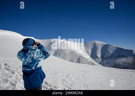 Un garçon de 10 ans regarde à travers des jumelles une belle montagne enneigée dans les Tatras occidentales de Baranec, le temps est hiver et beau Banque D'Images