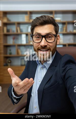Gros plan vertical d'un jeune homme dans un costume de travail et des lunettes assis dans le bureau, parlant en ligne et faisant des gestes vers l'appareil photo de l'appareil. Banque D'Images