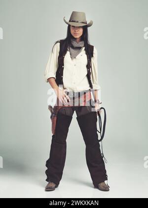 Femme, cowgirl et portrait en studio pour costume de mode avec confiance sur fond gris, arme ou hors-la-loi. Femme personne, fouet et chapeau avec Banque D'Images