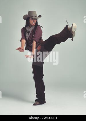 WESTERN, femme et cow-boy en studio avec mode, jambe haute et geste pour se battre sur fond gris. Femme personne, vintage et hors-la-loi avec des bottes pour Banque D'Images