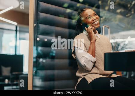 Jeune femme d'affaires noire prospère assise dans un espace de coworking, parlant sur un smartphone avec un sourire heureux. Elle incarne le professionnalisme et la distance w Banque D'Images