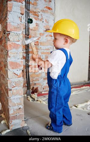 Toute la longueur de l'enfant travailleur de la construction debout par le mur de briques et tenant le marteau. Enfant portant un casque de sécurité et des combinaisons de travail tout en utilisant l'instrument de construction à la maison pendant la rénovation. Banque D'Images