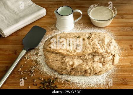 Pâte de levain crue sur une planche de bois avec des graines de citrouille et des ustensiles de cuisine Banque D'Images