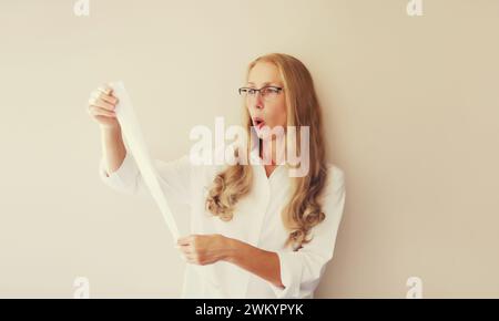 Choqué et surpris femme d'âge moyen caucasien dans des lunettes semble étonné par l'étiquette de prix élevé sur le long reçu de papier Banque D'Images