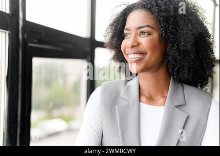 Une femme d'affaires afro-américaine rayonnante regarde par la fenêtre avec un regard optimiste, reflétant une attitude avant-gardiste dans un cadre de bureau contemporain et bien éclairé. Banque D'Images