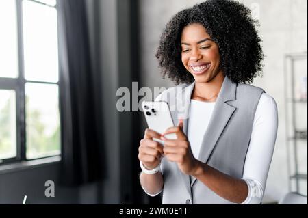 Une femme d'affaires afro-américaine est capturée dans un moment de joie franc, naviguant sur son smartphone près de la fenêtre, baignée de lumière naturelle qui souligne sa présence confiante et technophile. Banque D'Images