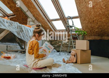 Fille peignant dans le livre assis près de la fenêtre de puits de lumière à la maison Banque D'Images