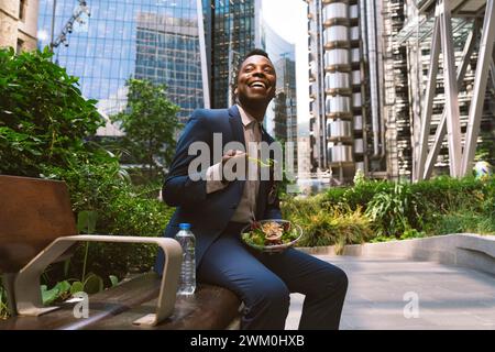 Heureux homme d'affaires assis avec un bol de salade sur le banc au parc de bureaux Banque D'Images