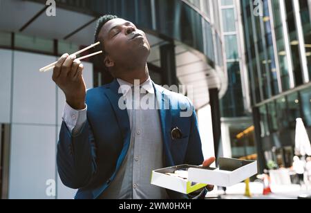 Homme d'affaires avec les yeux fermés appréciant des sushis devant les bâtiments Banque D'Images