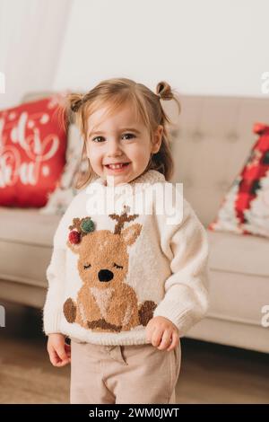 Fille mignonne souriante portant un pull de Noël près du canapé à la maison Banque D'Images