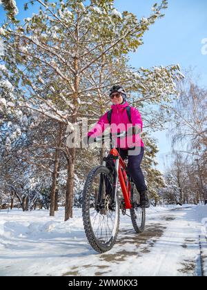 Aventure cycliste hivernale. Une fille en veste rose parmi les arbres enneigés fait du vélo le long d'un chemin dans le parc Banque D'Images
