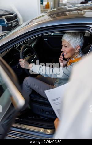 Femme senior souriante parlant sur téléphone intelligent dans la voiture à la salle d'exposition Banque D'Images