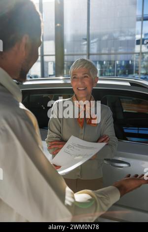 Femme senior heureuse avec vendeur tenant le document et montrant la voiture à la salle d'exposition Banque D'Images