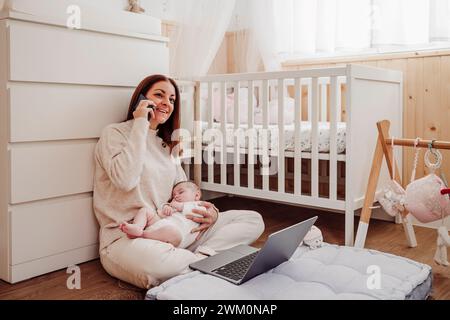 Souriant freelance tenant bébé fille et parlant sur téléphone intelligent au bureau à domicile Banque D'Images