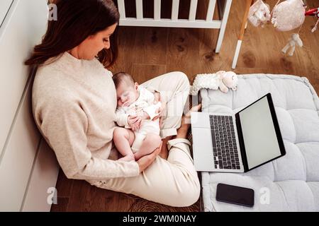 Mère prenant soin de bébé fille et travaillant avec ordinateur portable au bureau à domicile Banque D'Images