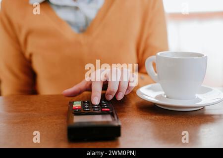 Femme entrant PIN sur le lecteur de carte de crédit près de tasse à café à table Banque D'Images