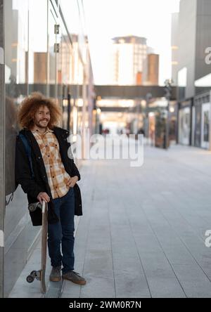 Homme heureux penché avec skateboard sur le mur de verre Banque D'Images