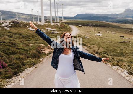 Espagne, Madrid, Portrait d'une femme enceinte debout avec les bras levés au milieu de la route s'étendant devant le parc éolien Banque D'Images