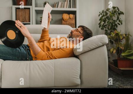Homme lisant du papier couché avec un vieux disque vinyle sur un canapé à la maison Banque D'Images