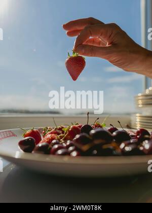 Main de femme tenant la fraise près de la fenêtre à la maison Banque D'Images