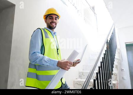 Architecte souriant portant un casque et debout sur les marches tenant l'ordinateur portable sur le site Banque D'Images