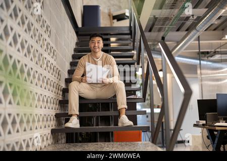 Souriant jeune homme d'affaires avec des documents assis sur l'escalier au bureau Banque D'Images