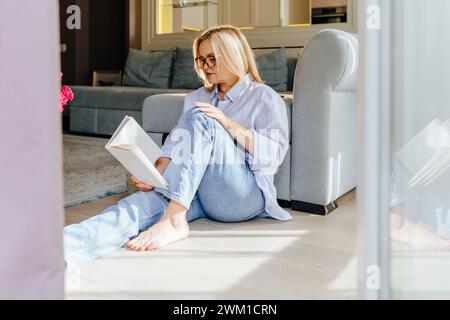 Mature femme de 55 ans bien soigné femme blonde avec des lunettes, habillé en Jean et chemise bleue en lin, assis sur le sol du salon, lisant a Banque D'Images