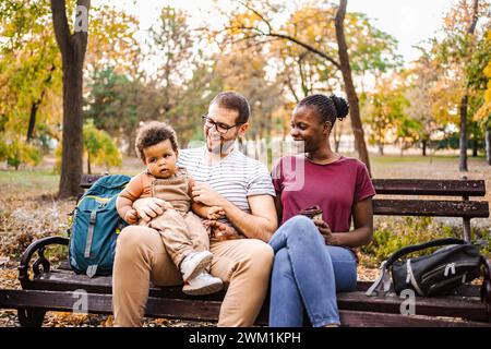 Un couple de races mixtes est assis sur un banc de parc avec leur fils. Banque D'Images