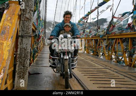 Motocycliste & enfant sur un pont couvert de drapeaux de prière. Le pont est au-dessus de la rivière Kyi Chu près de Lhassa dans la région autonome du Tibet en Chine. Banque D'Images