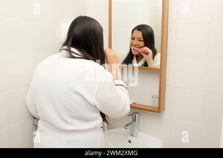 Latina femme de 40 ans se brosse et prend soin de ses dents pour prévenir les caries habillées en peignoir dans la salle de bain devant le miroir Banque D'Images