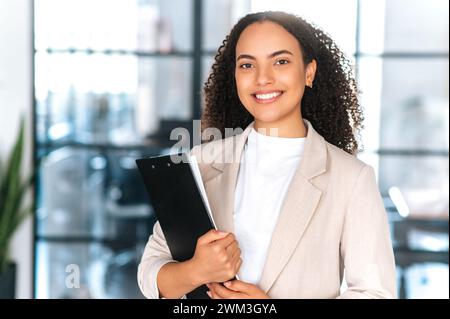 Portrait d'une belle femme d'affaires hispanique aux cheveux bouclés positive dans des vêtements élégants, secrétaire, femme d'affaires, tenant des documents tout en se tenant debout dans le bureau moderne, sourire à la caméra Banque D'Images