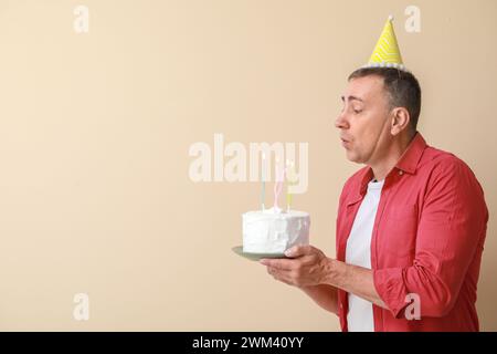 Homme mature avec gâteau d'anniversaire faisant souhait sur fond beige Banque D'Images