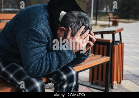 Sans-abri vieux homme caucasien âgé avec dépression tient la tête dans les mains tout en étant assis sur un banc de parc Banque D'Images