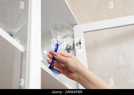 la main d'une femme sort un verre à martini de l'armoire. armoire de cuisine avec vaisselle. videz le verre à martini. alcoolisme féminin. verre à cocktail Banque D'Images