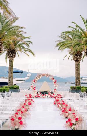 Des rangées de chaises bordent un chemin blanc décoré de fleurs rouges devant une arche de mariage ronde sur la jetée Banque D'Images