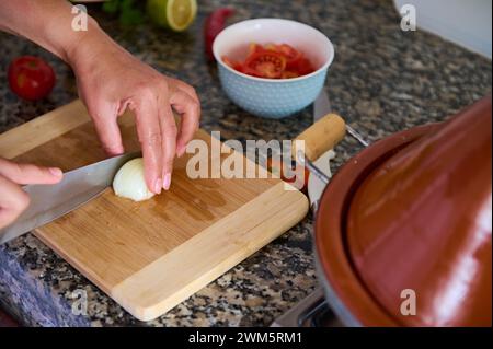 Vue rognée des mains d'un chef coupant l'oignon en deux moitiés sur une planche de bois, à l'aide d'un couteau de cuisine. Femme au foyer prépare le dîner dans la cuisine de la maison. C Banque D'Images