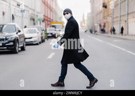 Homme masqué traversant avec confiance la rue urbaine avec le journal. Banque D'Images