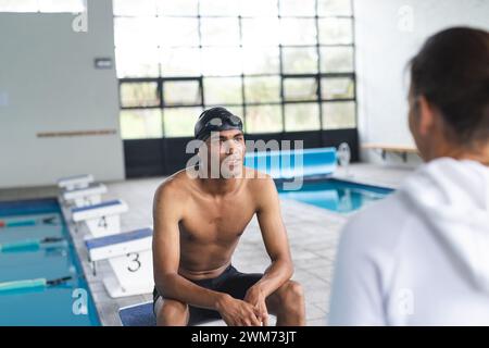 Jeune nageur athlète masculin biracial assis au bord de la piscine, s'engageant dans la conversation Banque D'Images