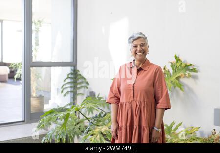 Femme biraciale senior se tient souriante dans une maison lumineuse, avec espace de copie Banque D'Images