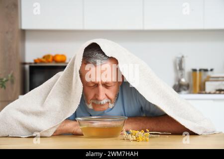 Homme mûr avec serviette faisant inhalation de vapeur à table dans la cuisine Banque D'Images