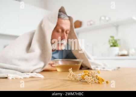 Homme mûr avec serviette faisant inhalation de vapeur à table dans la cuisine Banque D'Images