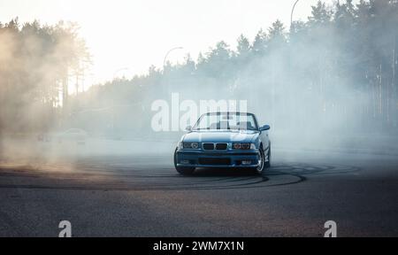 Blue BMW M3 (E36 ) sur un grand parking couvert de fumée de pneu. Vue de face du cabriolet sur terrain pavé près de la forêt. Banque D'Images