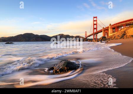 Vagues lavant sur la plage avec des rochers éparpillés au premier plan et de beaux tons rouges du Golden Gate Bridge dans la distance éclairée par le soleil couchant Banque D'Images