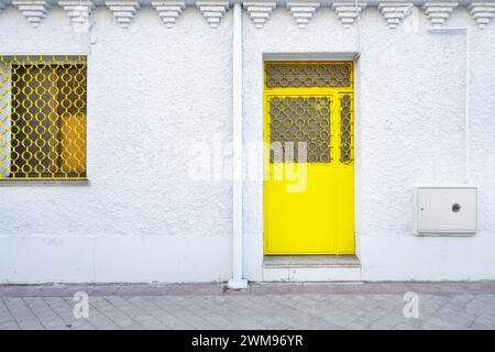 Porte jaune avec serrure de sécurité et barres métalliques sur les fenêtres sur une façade en ciment blanc dans un local au rez-de-chaussée Banque D'Images